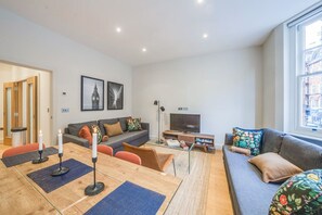 Uma sala de estar moderna com um sofá, uma mesa de centro e uma televisão. Há uma porta que dá para outro cômodo e uma janela com vista.