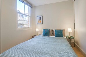Quarto com cama, duas mesas de cabeceira com luminárias, janela com vista para edifícios e um quadro na parede.