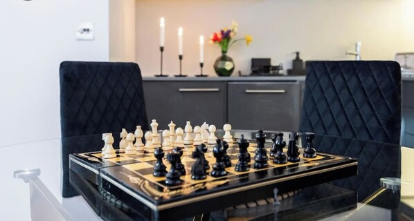A chessboard on a glass table with a modern interior, including dark upholstered chairs and a neutral-toned cabinet in the background. - Luxury 2BR 2Bath Flat | Walk to Tube | West Ken (London)
