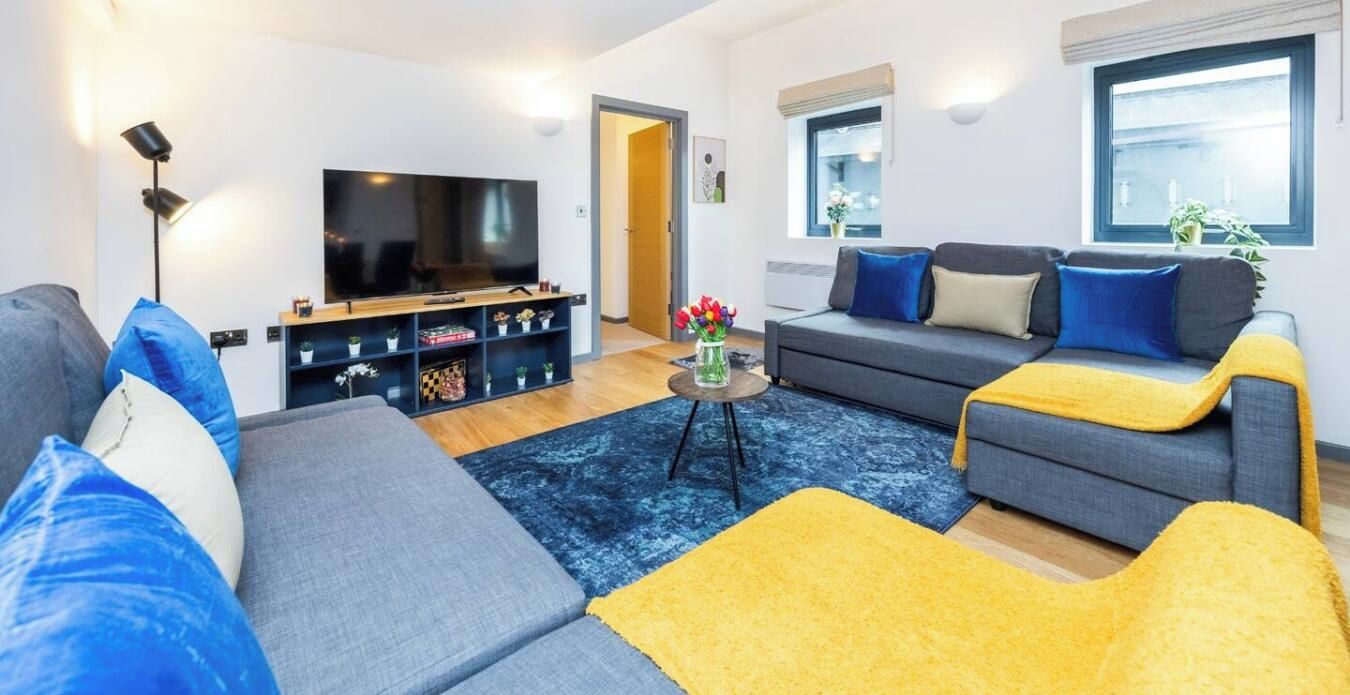 Uma sala de estar moderna com um sofá cinzento, um banquinho amarelo, uma TV de tela plana e uma mesinha de centro com objetos decorativos.