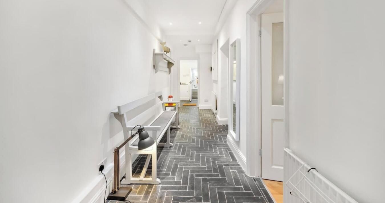 A narrow hallway with a herringbone floor pattern, white walls, and a bench with a lamp.