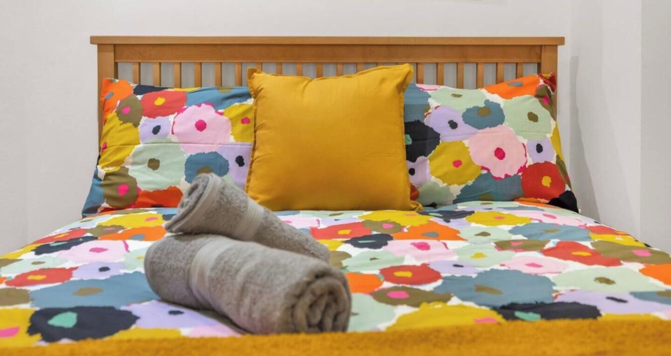 Ein Bett mit einer bunten, blumigen Tagesdecke, einem gelben Kopfkissen und einem zusammengerollten Handtuch.