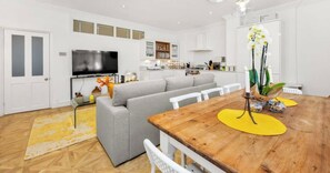 A modern living room with a sofa, a wooden dining table, and a kitchen in the background. - Victorian 3BR | 1-Min Tube | Work Ready (Greater London)
