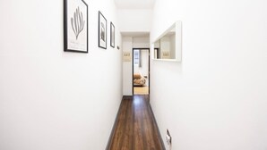 Een gang met kunstwerken in lijsten aan de wanden, een houten vloer en een deur naar een kamer met een bed.