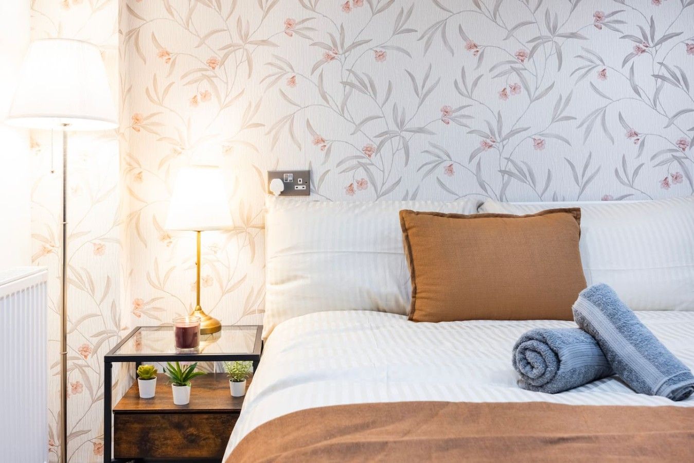Una cama bien hecha con una almohada marrón y toallas enrolladas, una mesita de noche con una lámpara y pequeñas plantas, y una pared con estampado floral.