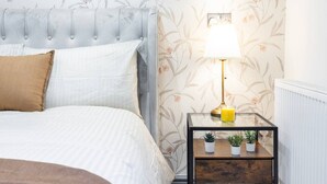 Een slaapkamer met een geborduurde hoofdbord, een nachtkastje met een lamp en twee potplanten.