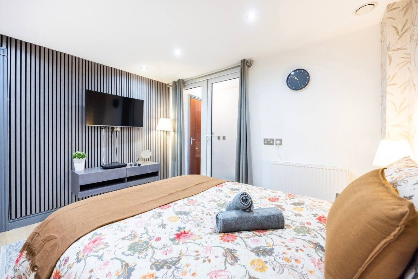 Une chambre moderne avec un téléviseur à écran plat fixé au mur, une table de chevet avec une lampe, un lit avec une literie à motifs floraux et une horloge au mur.