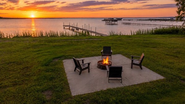 A grassy outdoor area with a fire pit and chairs, overlooking a body of water with docks.