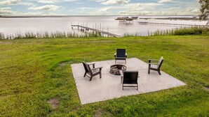 An outdoor seating area with chairs and a fire pit on a concrete slab, overlooking a body of water with docks and boats. - The Loon on North Long (Brainerd)