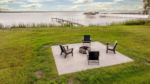Een terras buiten met stoelen en een vuurkorf op een betonnen plaat, met uitzicht op een waterpartij met steigers en boten.