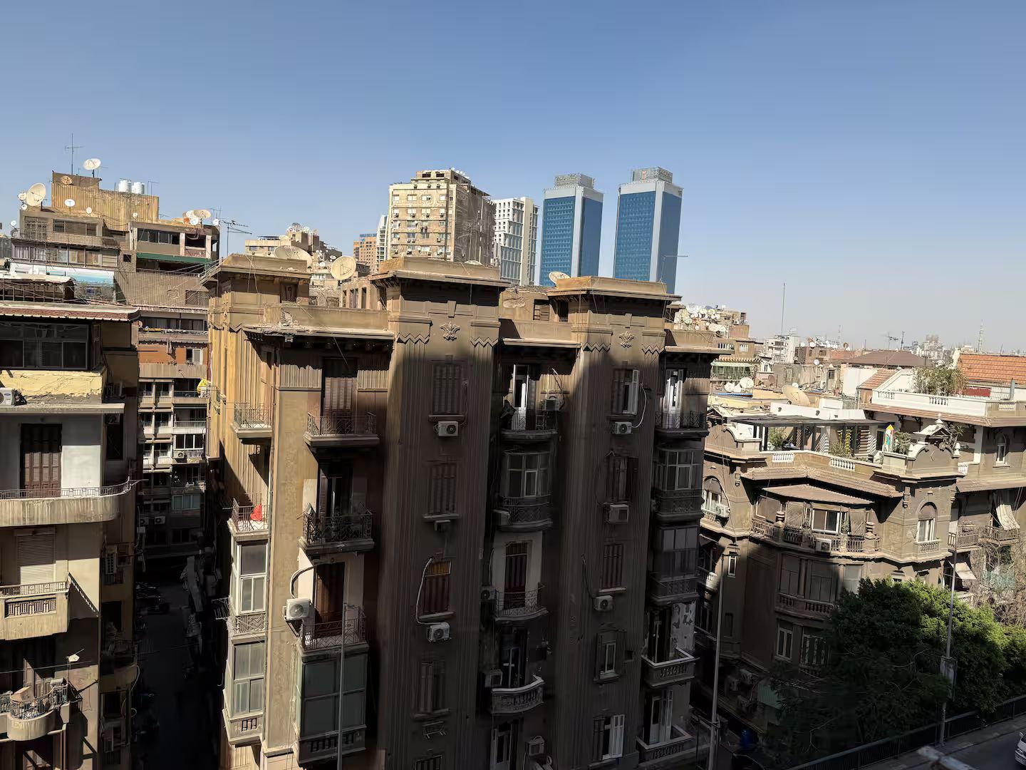 A multi-story building with balconies and satellite dishes, surrounded by other buildings and a clear sky.