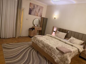 Ein Schlafzimmer mit einem Bett, einem Schminktisch, einem Spiegel, einer Wandleuchte und einem gemusterten Teppich.