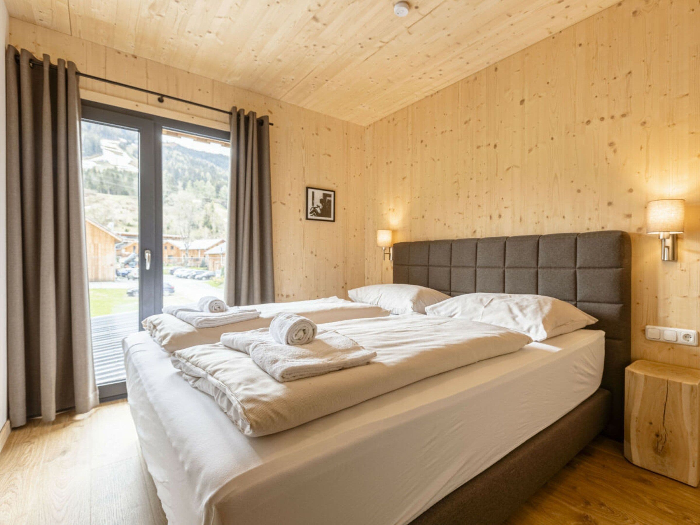 A hotel room with a wooden interior, a bed with white linens, a view through a glass door, and a bedside lamp.