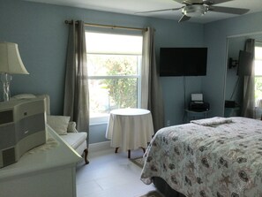 Habitación de hotel con cama, escritorio con lámpara, televisión en la pared, ventana con cortinas y ventilador de techo.