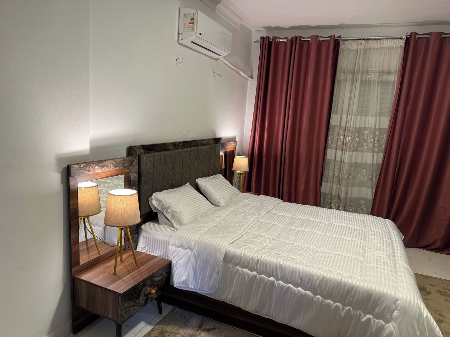 A hotel room with a bed, nightstands, lamps, an air conditioning unit, and a window with curtains.