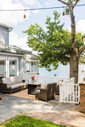 A covered outdoor seating area with a view of a lake, a large tree, and a house with a porch. - Bloom + Shore: Charming Lakefront Cottage on Conesus Lake (Geneseo)