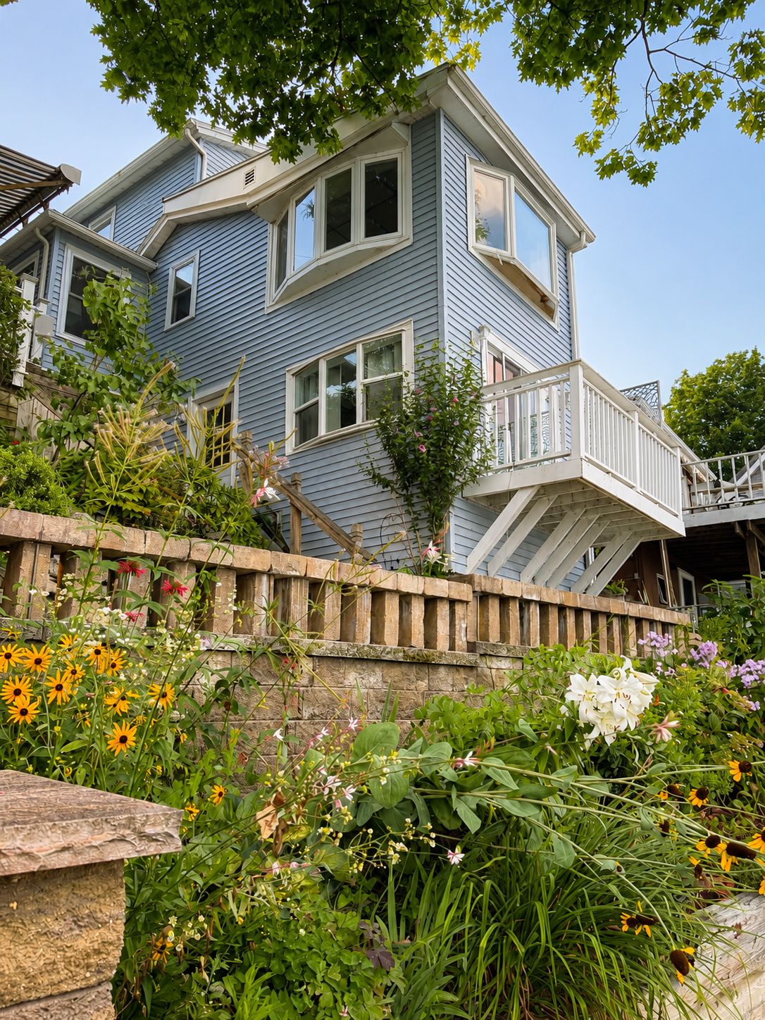 Une maison à deux étages, dotée d’un mur de soutènement en pierre et d’une terrasse en bois.