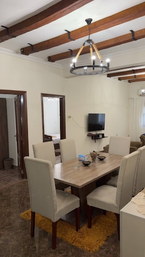 Comedor con mesa de madera, seis sillas, una lámpara de araña, televisor y alfombra amarilla.