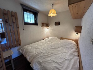A single bed with a quilted white duvet, a wooden headboard, a small window with blinds, and a wall-mounted light fixture. - NEW - NANT SOLEIL  A14 – Apartment with view, close to the village – Garage (La Clusaz)