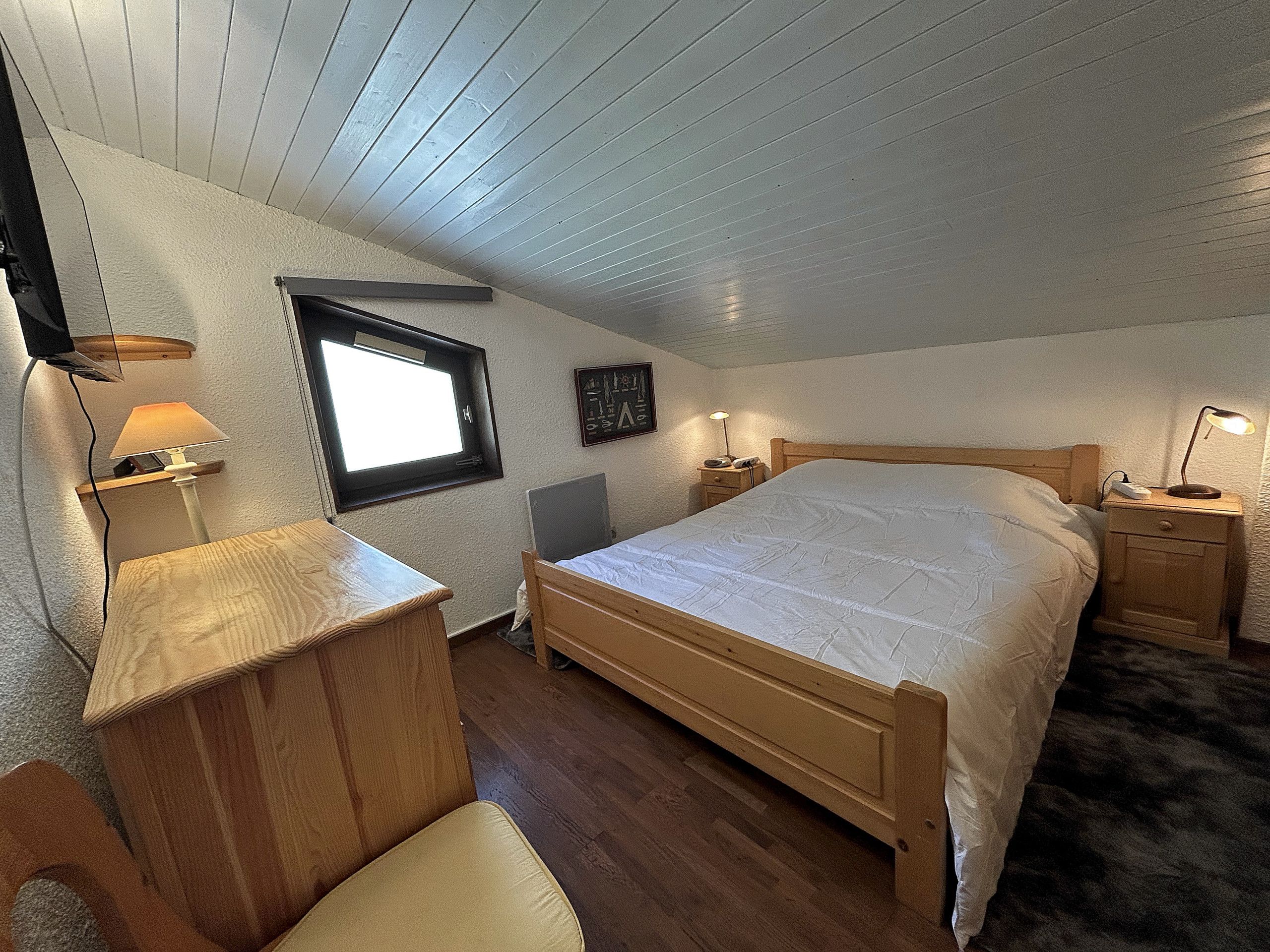 A bedroom with a wooden bed, a desk with a lamp, a window, and a wall-mounted shelf.