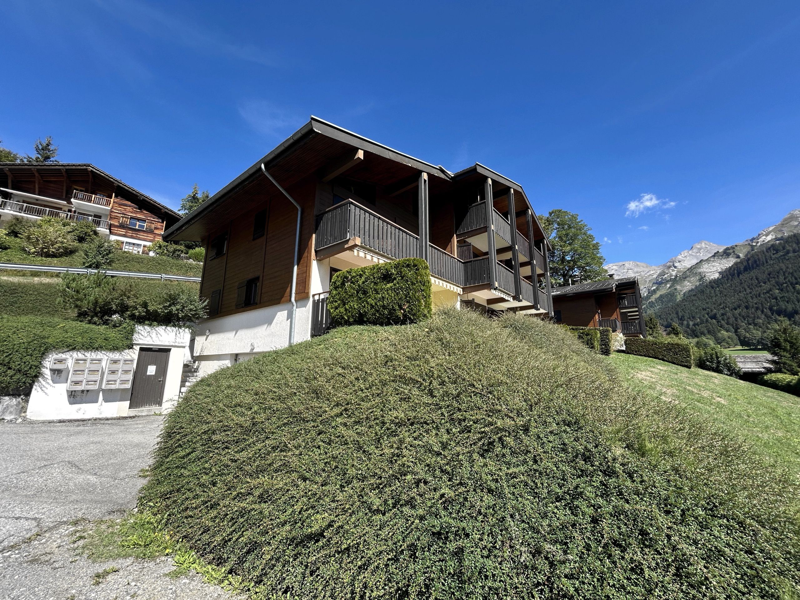 A chalet-style building with a balcony, situated on a hillside with a mountain backdrop.