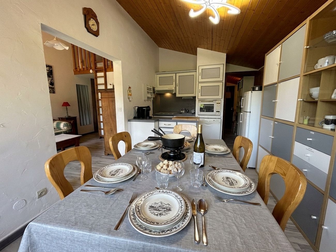 Ein Essbereich mit einem Tisch für sechs Personen, Holzkstühlen, einer Kronleuchter und einer Küche im Hintergrund.