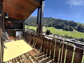 A balcony with a wooden railing, a yellow table, and a view of a green valley with trees and houses. - NEW - NANT SOLEIL  A14 – Apartment with view, close to the village – Garage (La Clusaz)