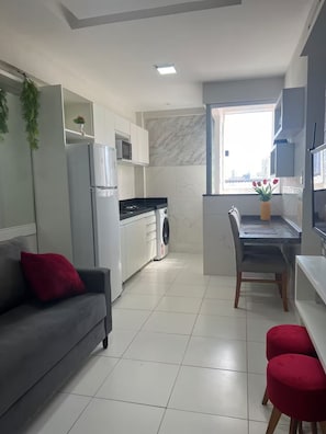 A modern kitchen with a refrigerator, microwave, and washing machine, a dining area with a table and chairs, and a gray sofa with red cushions. - 303 2-bedroom apartment 15 min center (Belém)