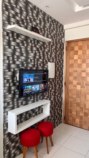 A modern living room with a flat-screen TV mounted on a textured wall, a white shelf, and two red stools. - 303 2-bedroom apartment 15 min center (Belém)