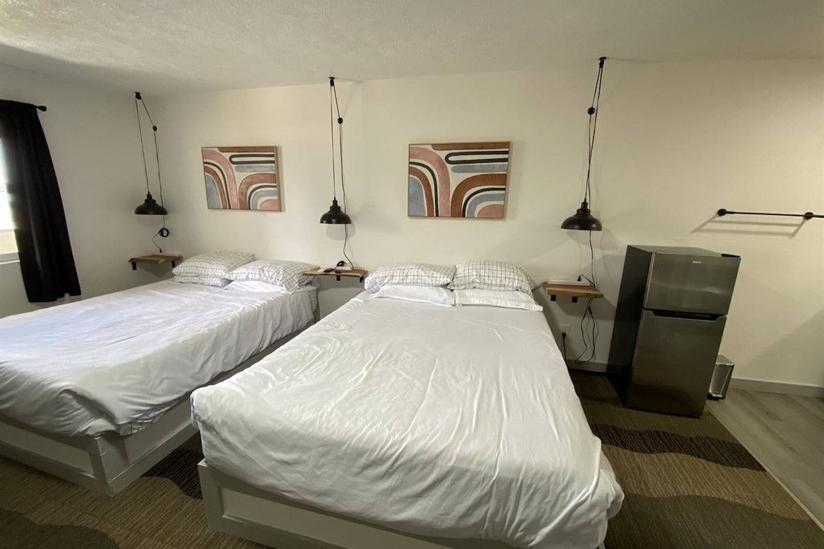 A hotel room with two single beds, a refrigerator, bedside tables, lamps, and abstract wall art.