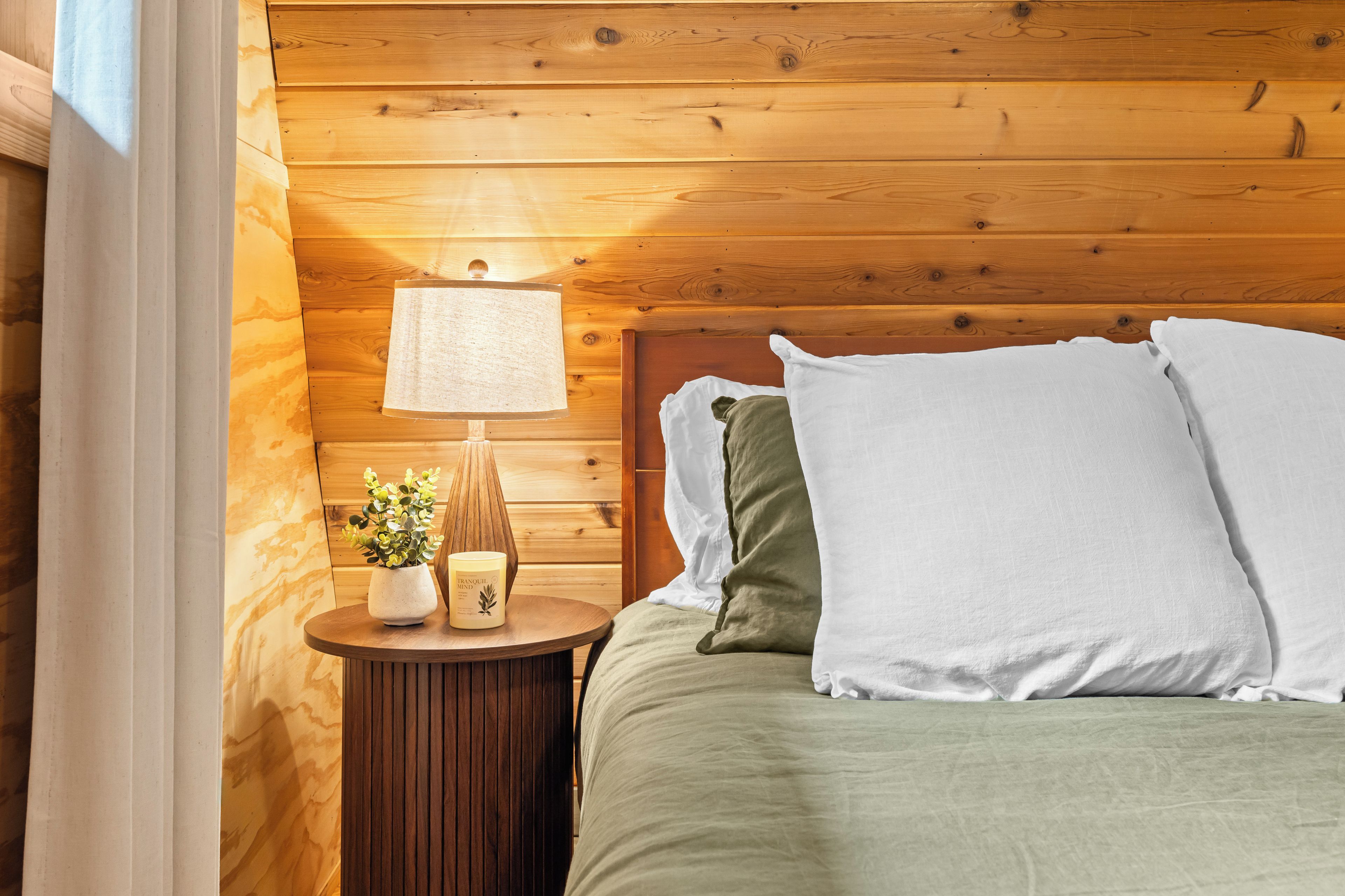 Una habitación con paneles de madera, una cama, almohadas, una mesita de noche con una lámpara y una planta pequeña.