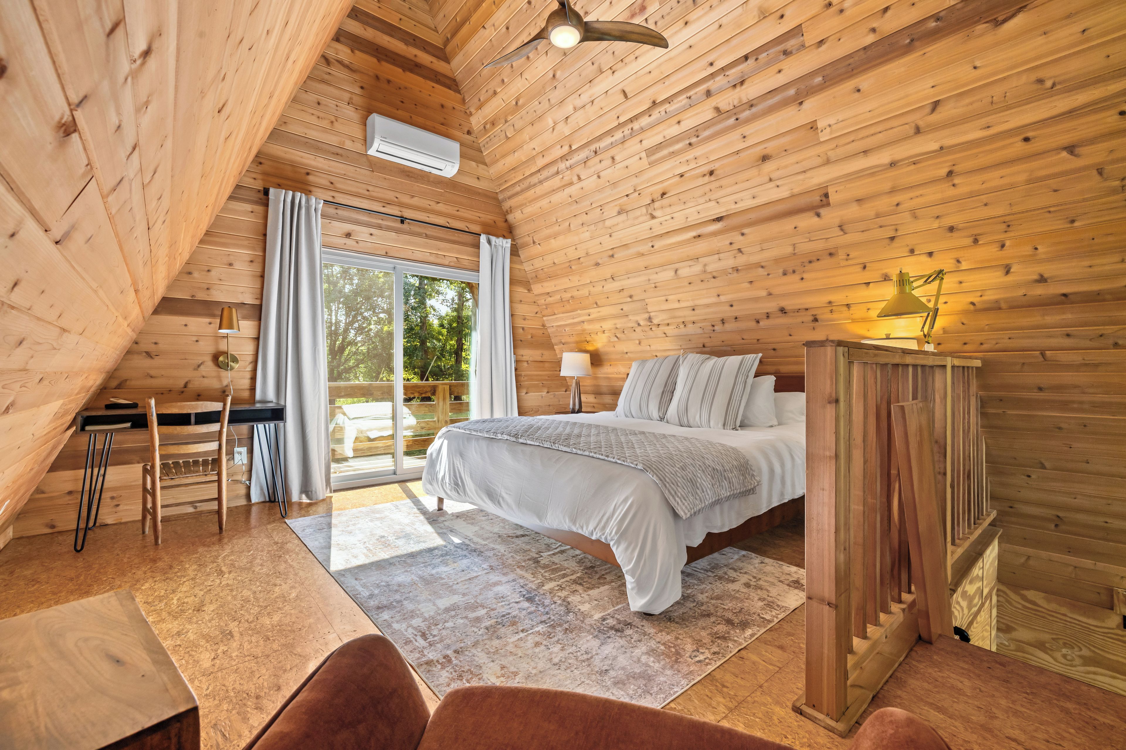 Un dormitorio con cama, escritorio, silla, lámpara y vista al exterior.