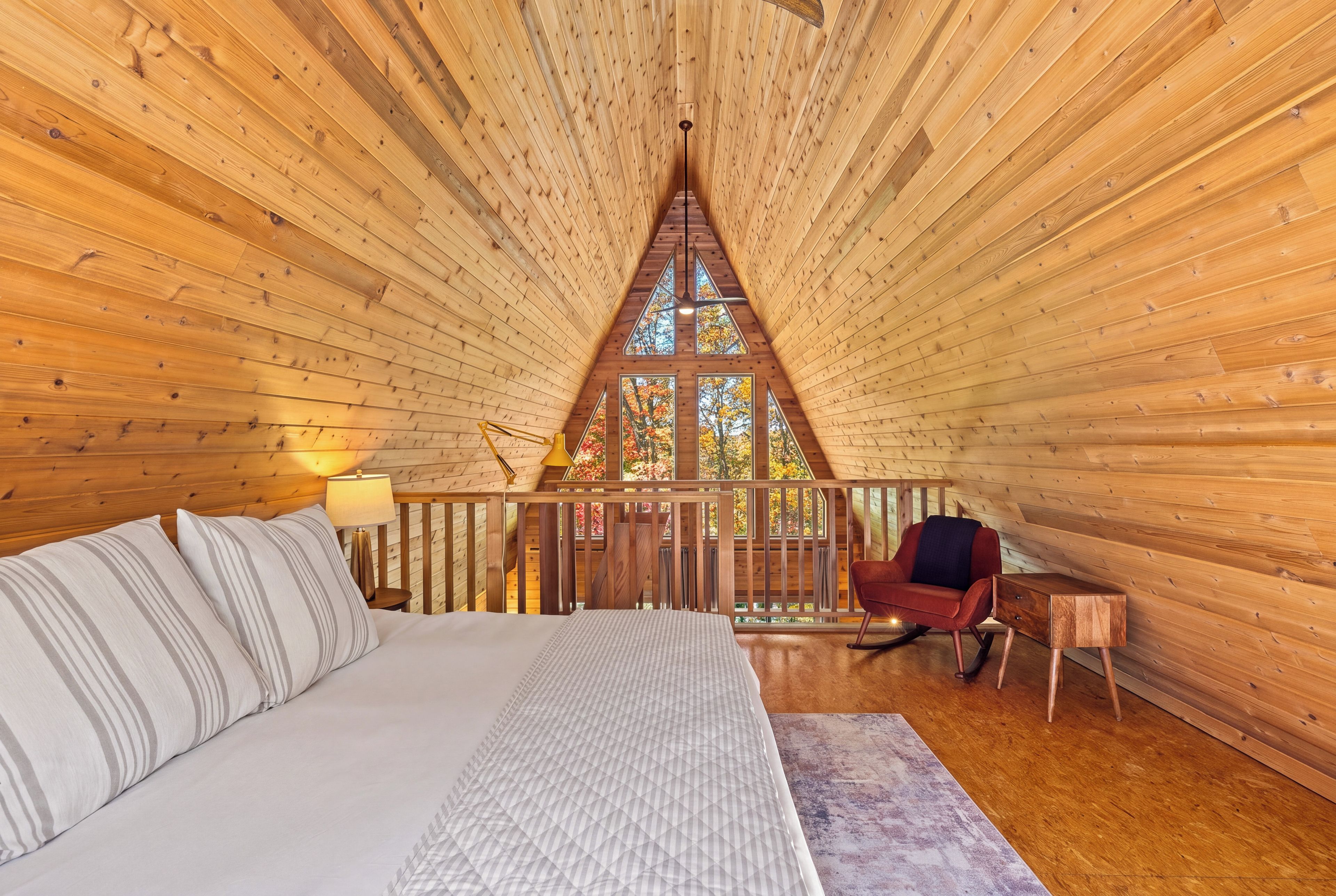 A cozy bedroom with a bed, a nightstand, a lamp, a rocking chair, and a view of autumn trees through a large window.