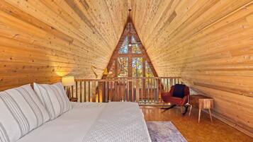 A cozy bedroom with a bed, a nightstand, a lamp, a rocking chair, and a view of autumn trees through a large window.