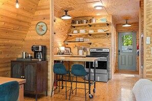 A compact kitchen with a coffee machine, a stove, and a sink. There are two bar stools and a wooden dining table.