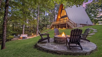 A lodge with a stone fire pit and Adirondack chairs, surrounded by a lush green landscape.