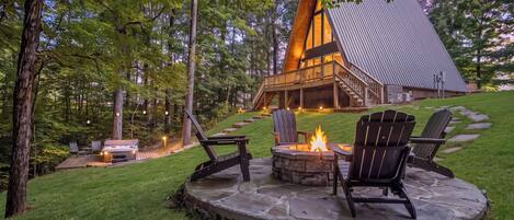 Una cabaña con un hogar de piedra y sillas Adirondack, rodeada de un paisaje verde exuberante.