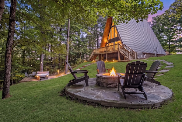 A lodge with a stone fire pit and Adirondack chairs, surrounded by a lush green landscape.