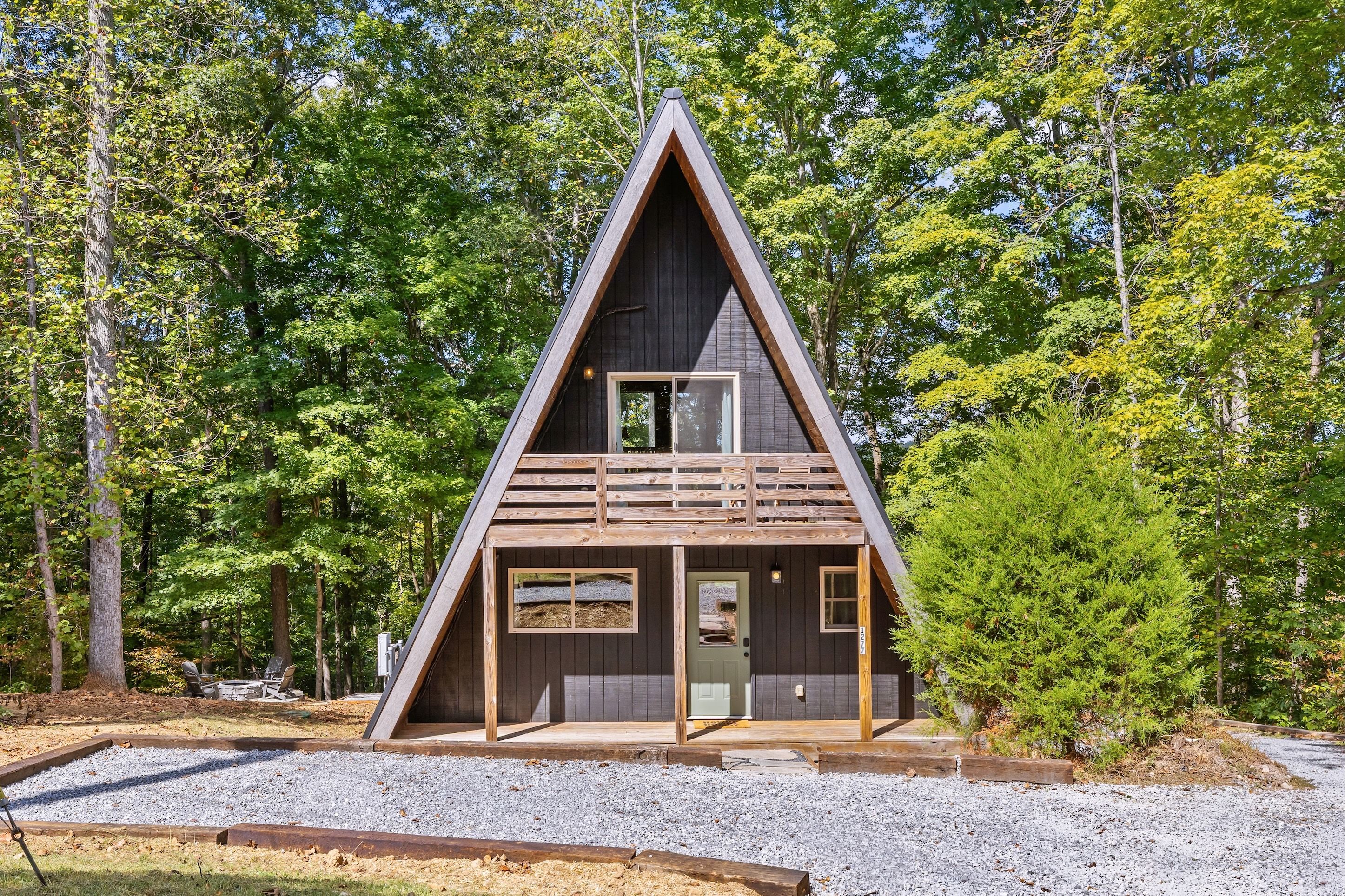 Una cabaña de madera triangular con un porche delantero y una pequeña terraza, rodeada de árboles.