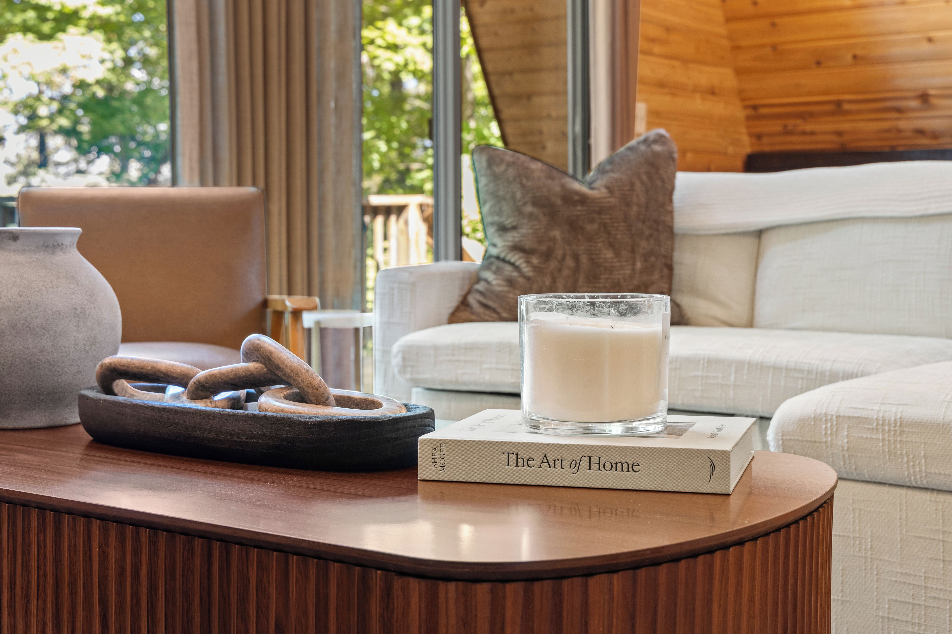 A wooden table with a book titled 'The Art of Home,' a glass candle, and a stoneware jug.