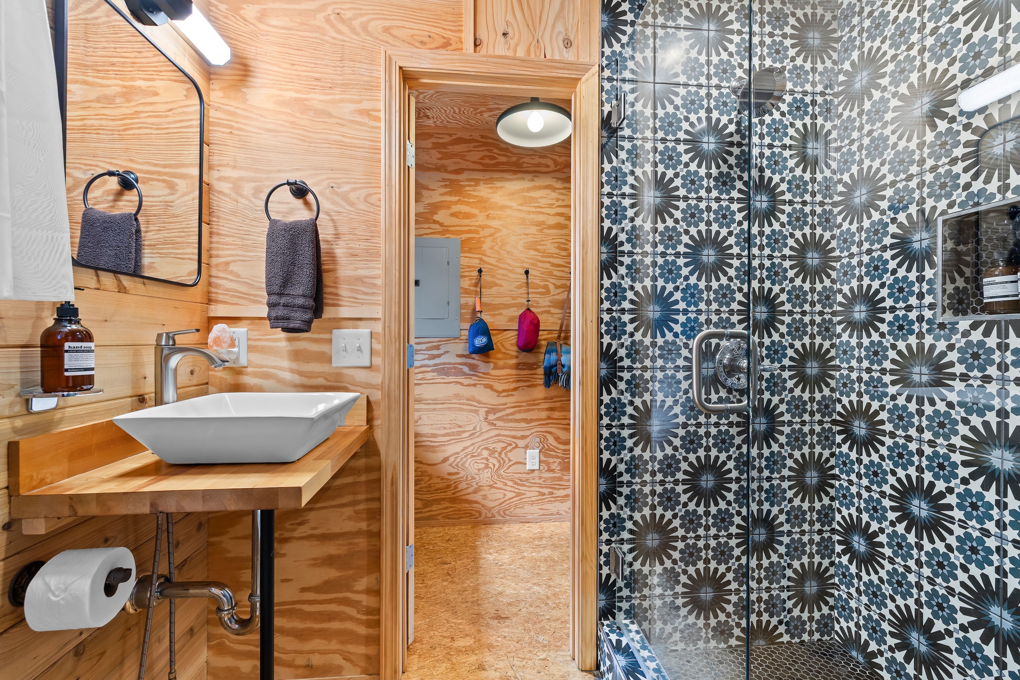 A bathroom with a wooden vanity, a white sink, a mirror, and a shower with patterned tiles.