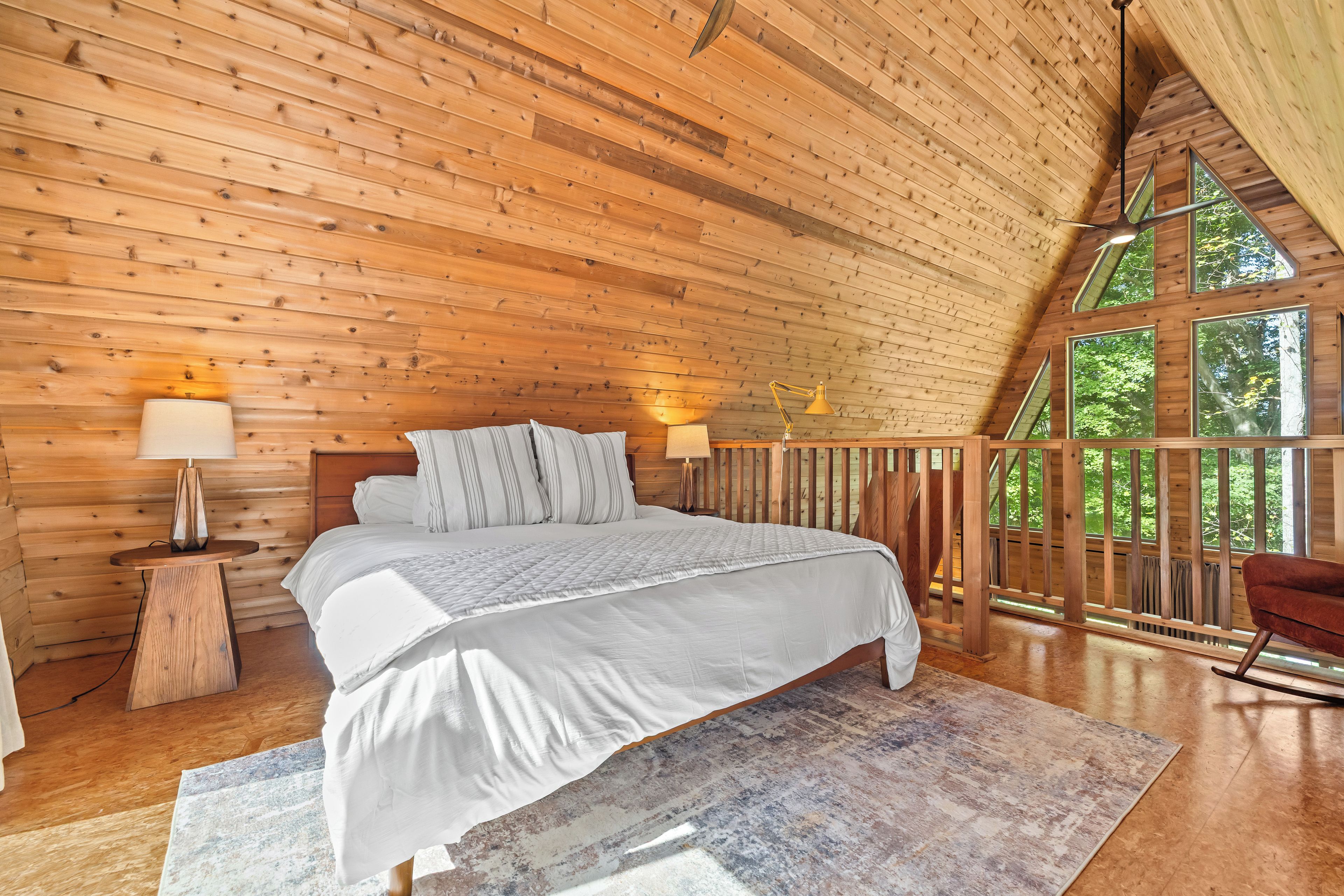A bedroom with a wooden ceiling, a bed with white bedding, two bedside lamps, and a view of greenery through large windows.