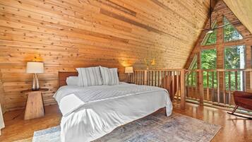A bedroom with a wooden ceiling, a bed with white bedding, two bedside lamps, and a view of greenery through large windows.
