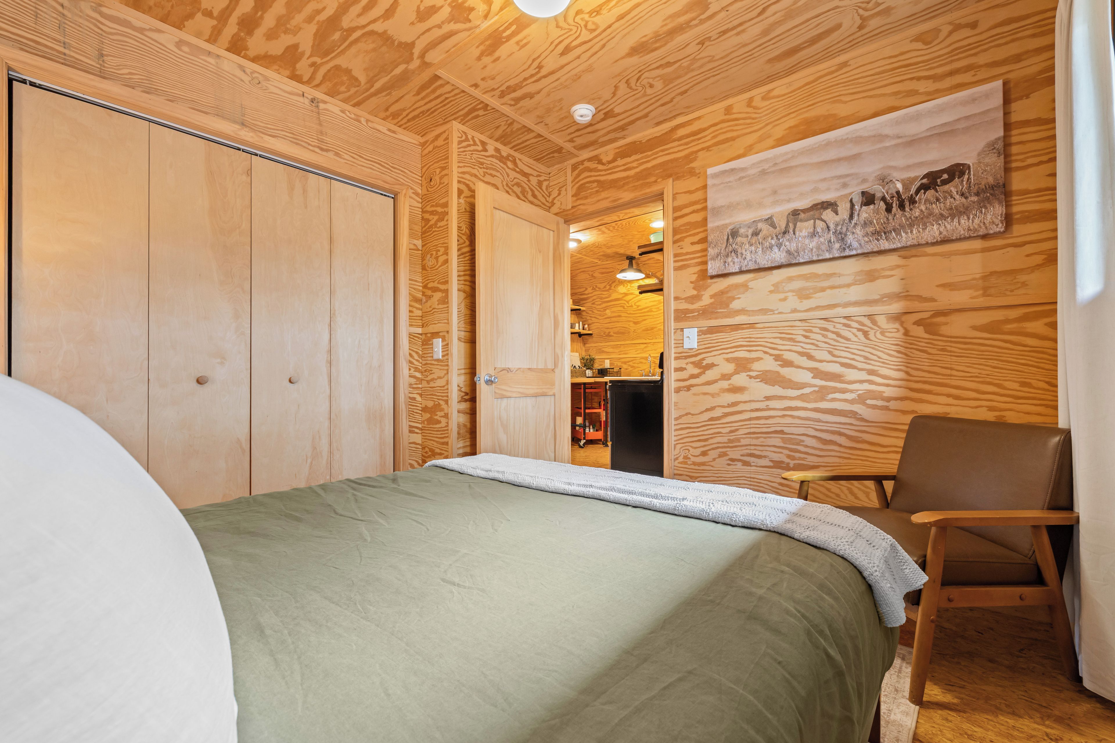 Een kamer met een bed, een stoel, houten wanden, een schilderij en een keukenhoek.
