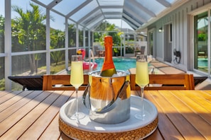 A table with a bottle of champagne and two glasses, a pool, and a covered patio area. - Heated Pool - 3BR Home in Marco Island - EV Charging (Marco Island)