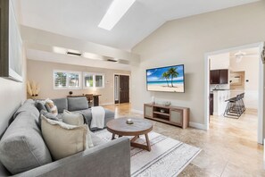 A spacious living room with a gray sofa, a coffee table, and a TV mounted on the wall. There is a kitchen visible through an open doorway. - Heated Pool - 3BR Home in Marco Island - EV Charging (Marco Island)