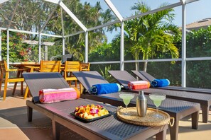 Un área de descanso al aire libre iluminada por el sol, con sillones, una mesa con una bandeja de frutas y bebidas, y vistas a exuberante follaje.