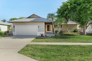 A single-story house with a garage, a well-maintained lawn, and a large tree in front. - Heated Pool - 3BR Home in Marco Island - EV Charging (Marco Island)