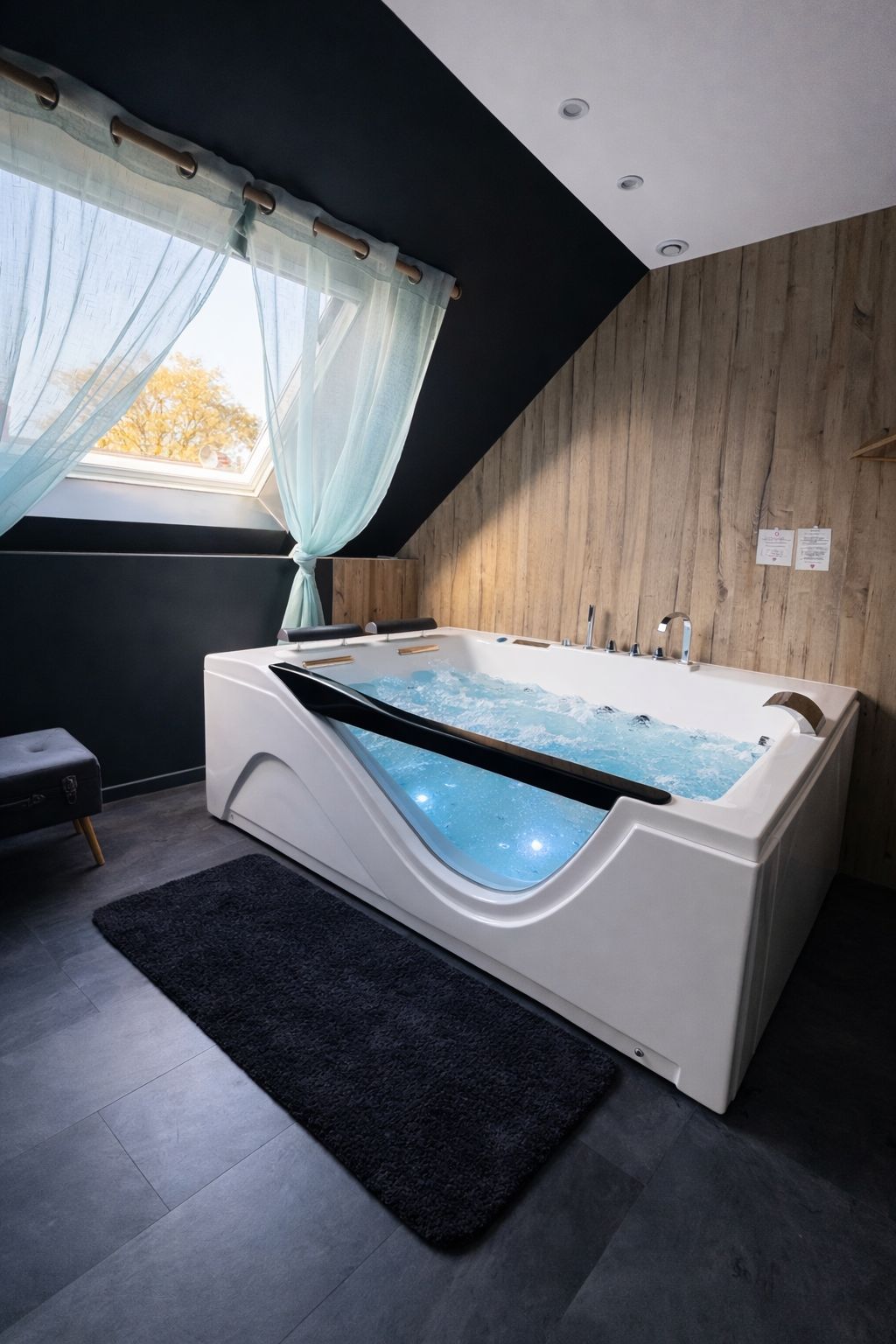 A modern bathroom with a large bathtub, a window with sheer curtains, and a wooden wall.