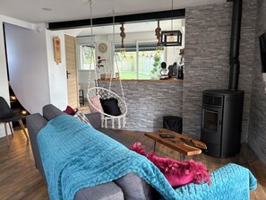 A modern living room with a sofa, a hanging chair, a fireplace, and a kitchen area visible through an open door. - Ker Love Rennes Nature maison 4 personnes (Laillé)